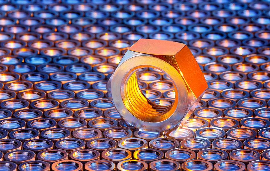 Sechskantmutter Eine große, glänzende Metallmutter steht auf einer Oberfläche aus vielen kleinen, runden metallischen Elementen. Das Licht reflektiert sich von der Mutter und den darunterliegenden Teilen, was zu einem schönen Farbspiel führt.