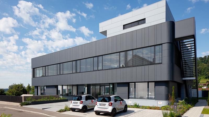 Modernes, mehrstöckiges Gebäude mit großen Fenstern und grauer Fassadenverkleidung. Vor dem Gebäude stehen zwei kleine Autos auf einem gepflasterten Parkplatz. Im Hintergrund sind Bäume und ein blauer Himmel mit einigen Wolken sichtbar.