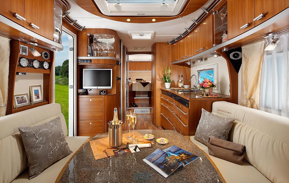 Luxuswohnwagen Ein modernes und elegantes Interieur eines Wohnmobils, das mit Holzverkleidungen, einem Esstisch und einer Sitzecke ausgestattet ist. Auf dem Tisch stehen Sektgläser, ein Kühlbehälter und Snacks. Im Hintergrund sieht man eine Küchenzeile und einen Fernseher. Große Fenster lassen Tageslicht herein.