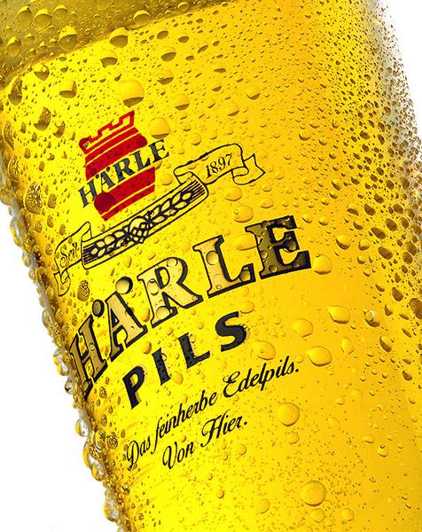 Bier Ein Glas mit goldenem Bier, das reich an Blasen ist. Auf dem Glas steht der Schriftzug "HARLE PILS" und darunter der Slogan "Das feinherbe Edelpils. Von Hier." Hintergrund ist unscharf.
