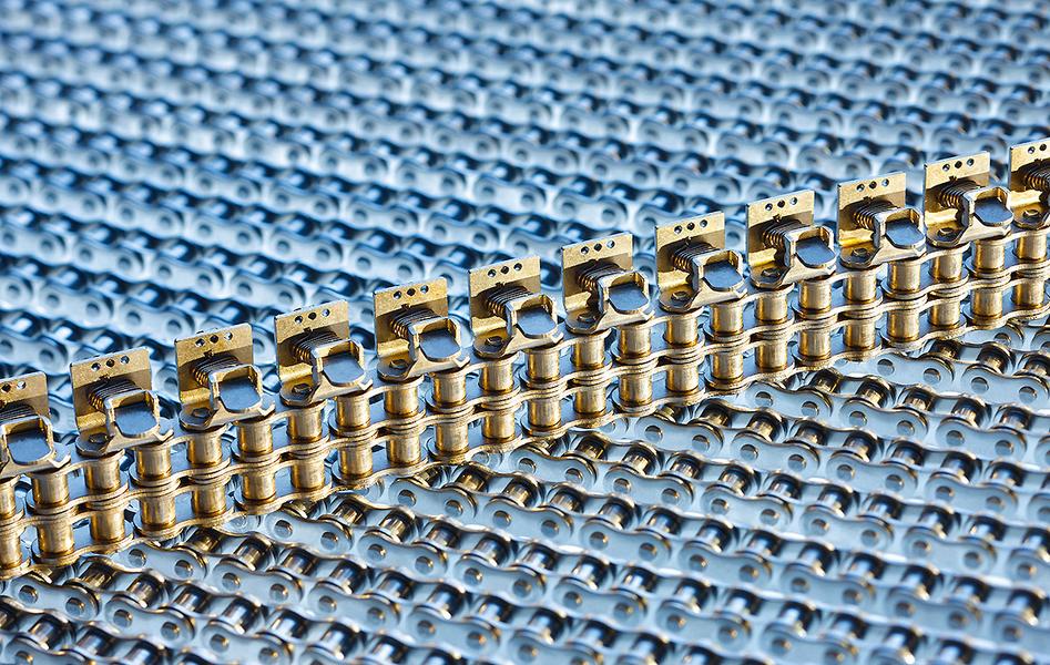 Antriebstechnik Ketten Eine Nahaufnahme von metallischen Kettenelementen auf einem blauen Hintergrund. Die Kette ist aus goldfarbenem Metall gefertigt und verläuft in gerader Linie. Die Struktur und Details der Kette sind gut sichtbar.