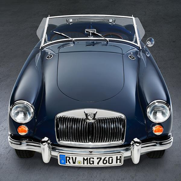 Ein blauer Oldtimer, ein MG, steht aus der Vogelperspektive auf einem glatten Untergrund. Die Motorhaube ist glänzend, und die Scheinwerfer leuchten. Die Windschutzscheibe ist klar, und das Fahrzeug hat keine Dachabdeckung, was einen offenen Eindruck vermittelt.