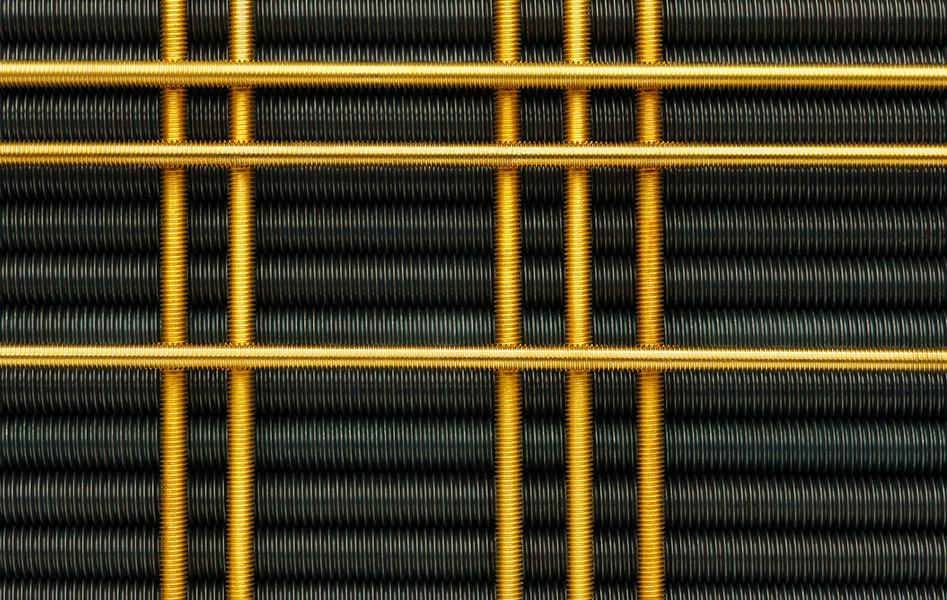 Nahaufnahme von Drahtgeflecht in schwarzer und goldener Farbe, das ein grafisches Muster bildet. Die schwarzen Drahtschlingen sind in Reihen angeordnet, während die goldenen Drähte quer verlaufen und ein strukturiertes, industrielles Design erzeugen.