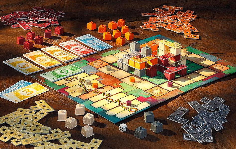 Gesellschaftsspiel Casa Grande Ein Tisch mit einem aufgeräumten Brettspiel. In der Mitte liegt das Spielbrett mit bunten Spielfiguren und Karten. Rund um das Brett sind Stapel aus farbigen Spielsteinen und Geldscheinen verteilt. Ein Würfel liegt ebenfalls auf dem Tisch.
