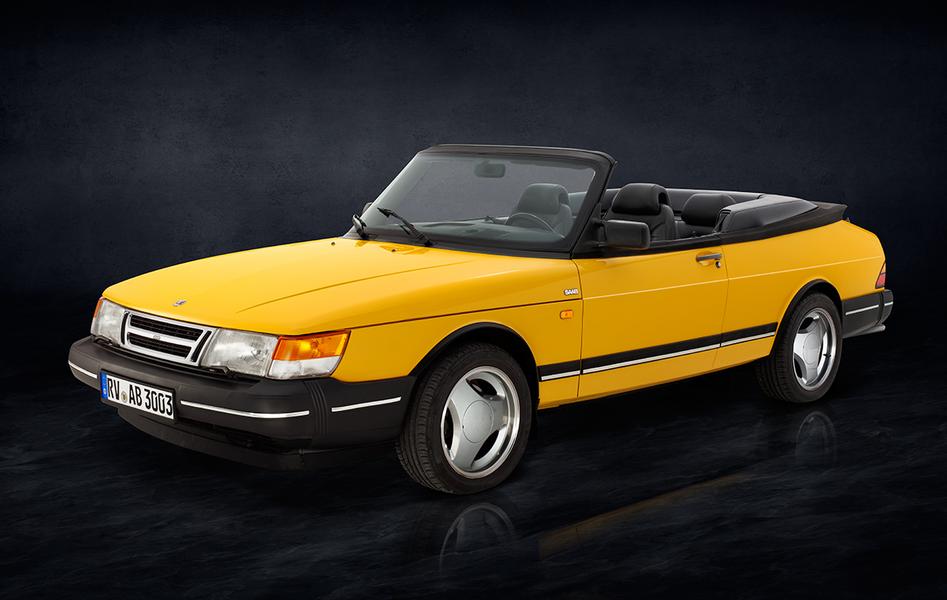 Ein gelber Sportwagen-Cabrio, Modell Saab, steht seitlich auf einer dunklen, einfarbigen Oberfläche. Das Auto hat schwarze Details und ein offenes Verdeck, sichtbar sind die beiden vorderen Sitze im Innenraum.