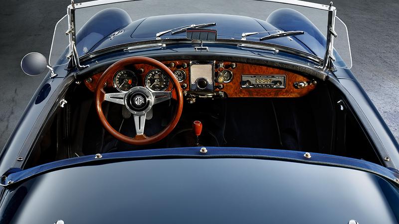 Eine Innenansicht eines klassischen Autos, das von oben fotografiert wurde. Der Blick zeigt das Lenkrad aus Holz, das Armaturenbrett mit verschiedenen Instrumenten und einem Radio. Die Sitze sind schwarz und der Fahrzeugboden ist gut sichtbar.