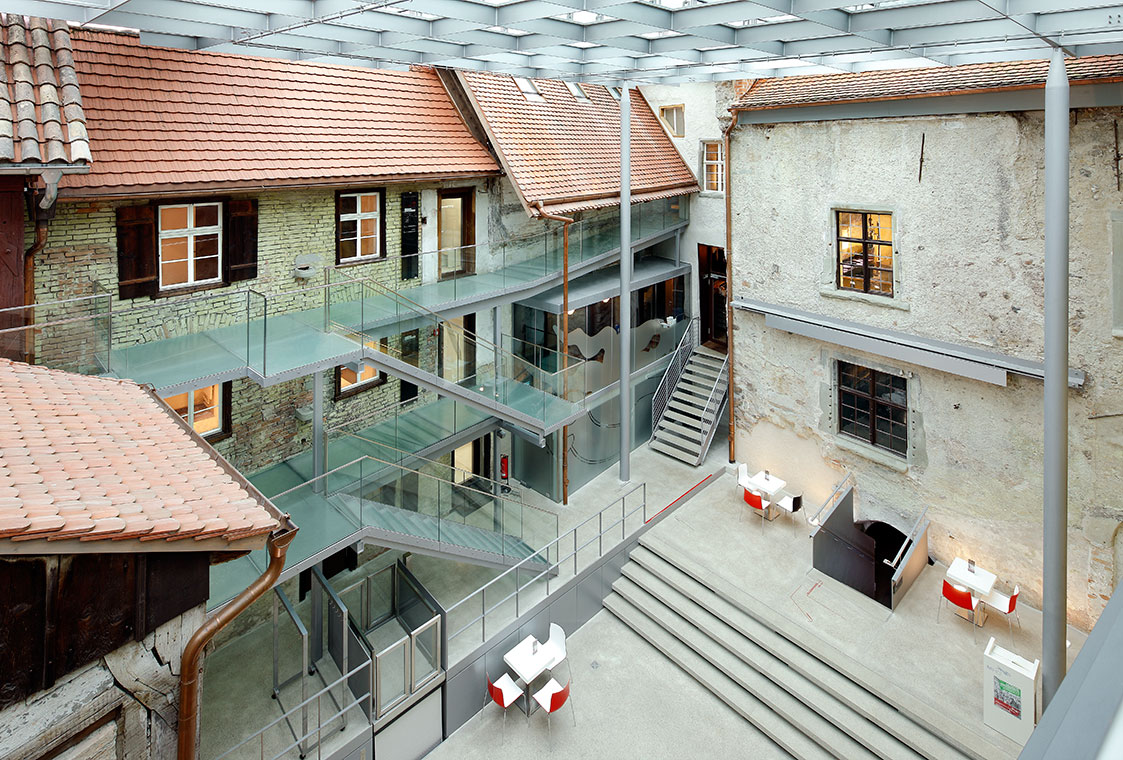 Innenansicht Humpis Museum Ein Überblick über ein modernes Atrium mit transparenten Glasböden und einer Kombination aus historischen Gebäuden. Die Umgebung ist hell erleuchtet, mit Treppen und Sitzbereichen. Die Architektur verbindet Alt und Neu in harmonischer Weise.