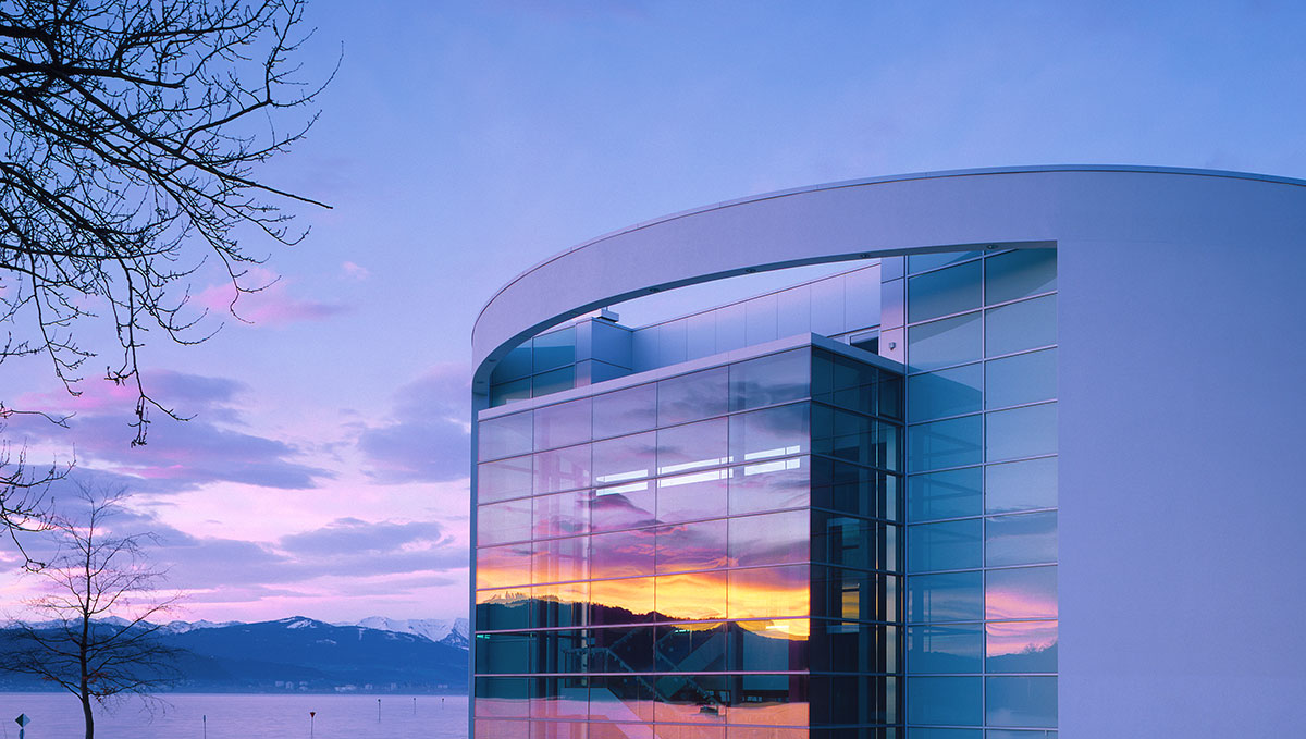 Moderne Gebäude mit großen Glasfenstern, reflektierend die Farben eines Sonnenuntergangs. Im Hintergrund sind Berge und ein ruhiger See sichtbar. Die Szene vermittelt eine friedliche Atmosphäre.