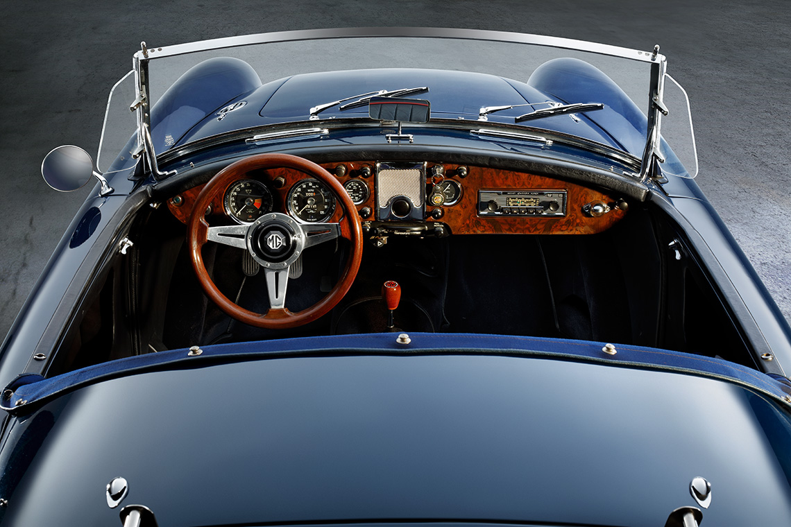Eine Innenansicht eines klassischen Autos, das von oben fotografiert wurde. Der Blick zeigt das Lenkrad aus Holz, das Armaturenbrett mit verschiedenen Instrumenten und einem Radio. Die Sitze sind schwarz und der Fahrzeugboden ist gut sichtbar.