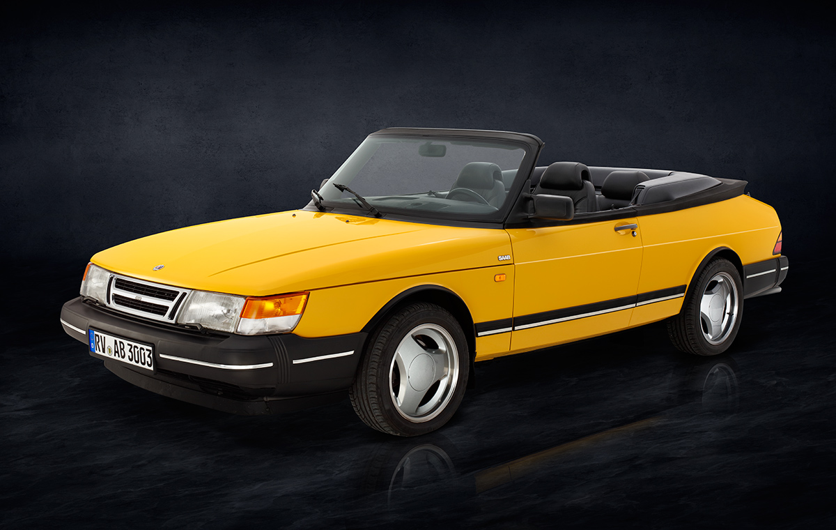 Ein gelber Sportwagen-Cabrio, Modell Saab, steht seitlich auf einer dunklen, einfarbigen Oberfläche. Das Auto hat schwarze Details und ein offenes Verdeck, sichtbar sind die beiden vorderen Sitze im Innenraum.