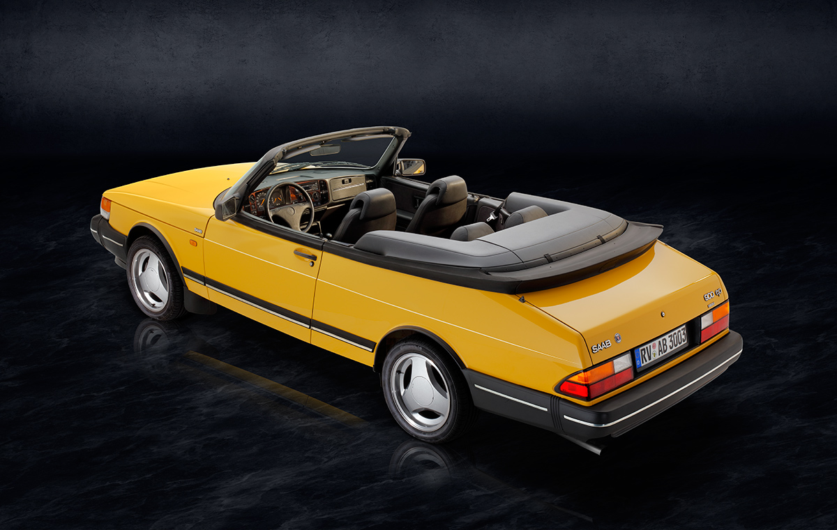 Ein gelbes Cabriolet mit schwarzem Verdeck steht vor einem dunklen Hintergrund. Die Rückansicht zeigt die charakteristischen Rundungen des Autos und die Designs der Felgen. Innenraumdetails sind teilweise sichtbar.