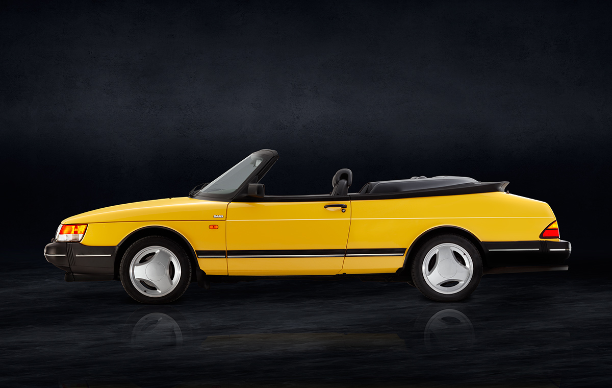 Ein gelber Cabriolet mit schwarzem Verdeck, seitlich abgebildet. Das Auto hat eine sportliche Silhouette und auffällige weiße Felgen. Der Hintergrund ist dunkel und hebt das Fahrzeug hervor.
