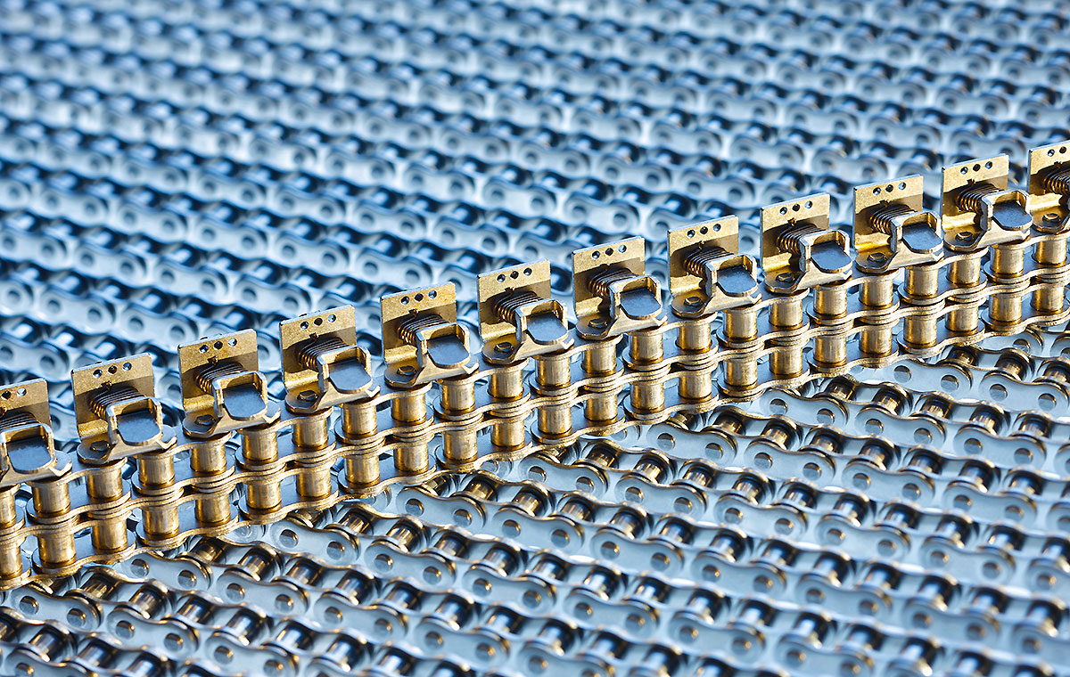 Antriebstechnik Ketten Eine Nahaufnahme von metallischen Kettenelementen auf einem blauen Hintergrund. Die Kette ist aus goldfarbenem Metall gefertigt und verläuft in gerader Linie. Die Struktur und Details der Kette sind gut sichtbar.