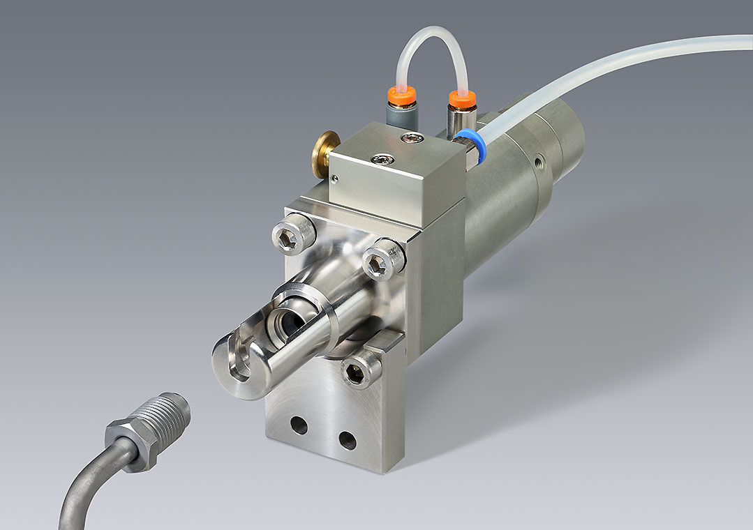 Anschluss Eine präzise Maschinenteileinheit, bestehend aus einem metallischen Gehäuse mit Anschlüssen und Schläuchen. Der Fokus liegt auf dem Ventilmechanismus, der für industrielle Anwendungen eingesetzt wird. Im Hintergrund ist eine neutrale graue Fläche.