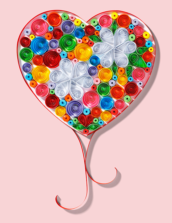 PaperArt Herz Ein herzförmiges Kunstwerk aus bunten Papierstreifen in unterschiedlichen Farben, verziert mit zwei weißen, blütenähnlichen Formen. Der Hintergrund ist rosa. Das Herz hat einen eleganten, geschwungenen Schwung am unteren Ende.