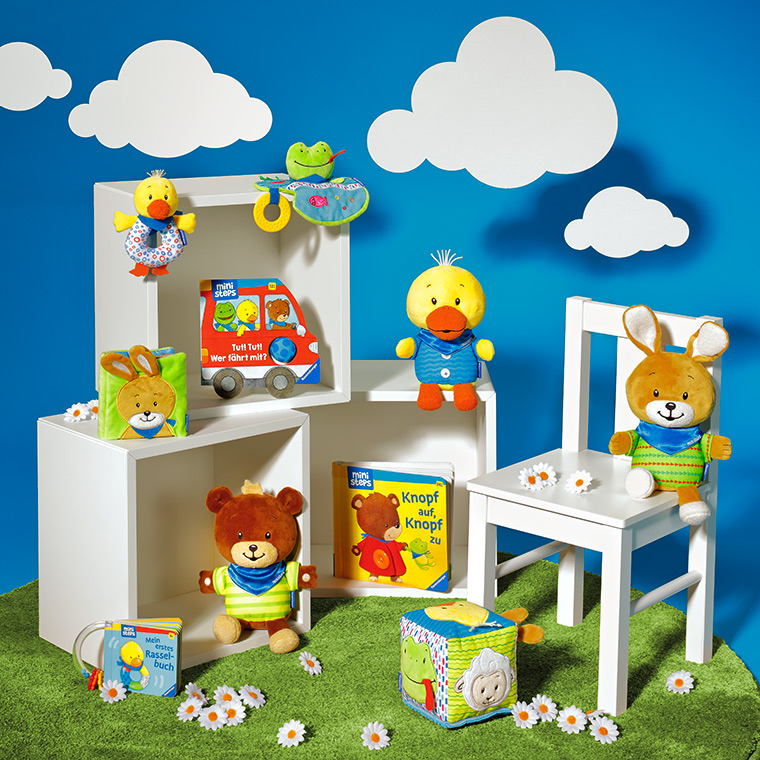 MS Neuheiten 2021 Bunte Spielzeuge und Plüschtiere auf einem grünen Teppich vor einer blauen Wand mit Wolken. Zu sehen sind ein Teddybär, eine Ente, ein Hase und weitere Spielartikel. Im Hintergrund stehen Bücher und ein Spielzeugauto.