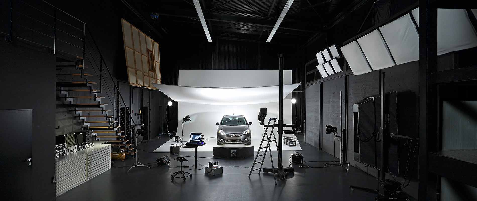 Ein modernes Fotostudio mit einem abgestellten Auto im Hintergrund. Die Szenerie ist gut beleuchtet, mit mehreren Lampen und einem weißen Hintergrund, der das Fahrzeug ins Licht rückt. An der Seite sind verschiedene Kamera- und Studioausrüstungen sichtbar.