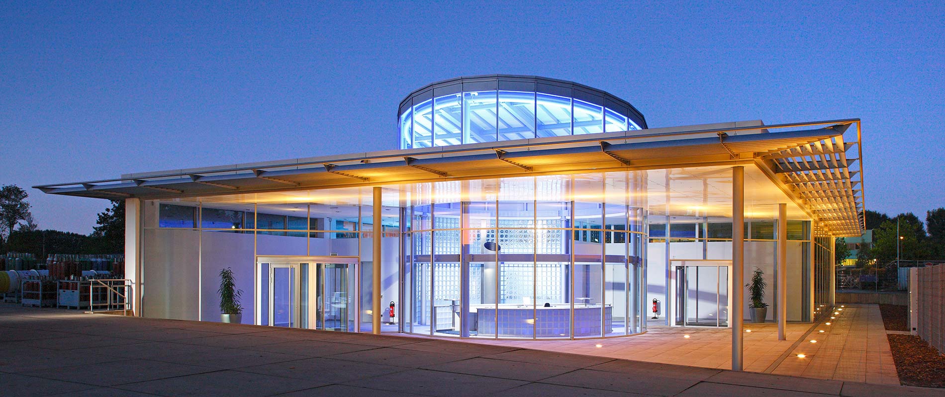 Moderne Architektur eines Gebäudes bei Dämmerung, das große Fensterfronten und einen runden Glaskuppelbereich aufweist. Das Gebäude ist beleuchtet und verfügt über einen großzügigen Eingangsbereich mit einer freundlichen Atmosphäre. Pflanzen sind in der Umgebung sichtbar.